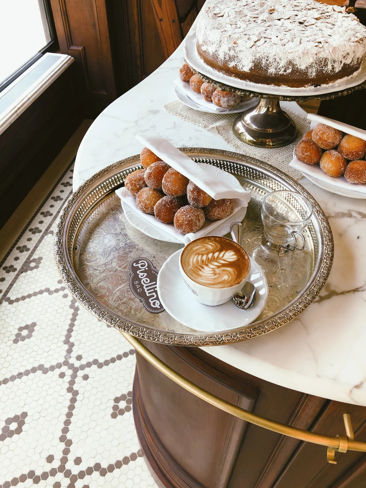 A branded Bar Pisellino cappuccino on a silver tray beside bomboloni and an almond cake